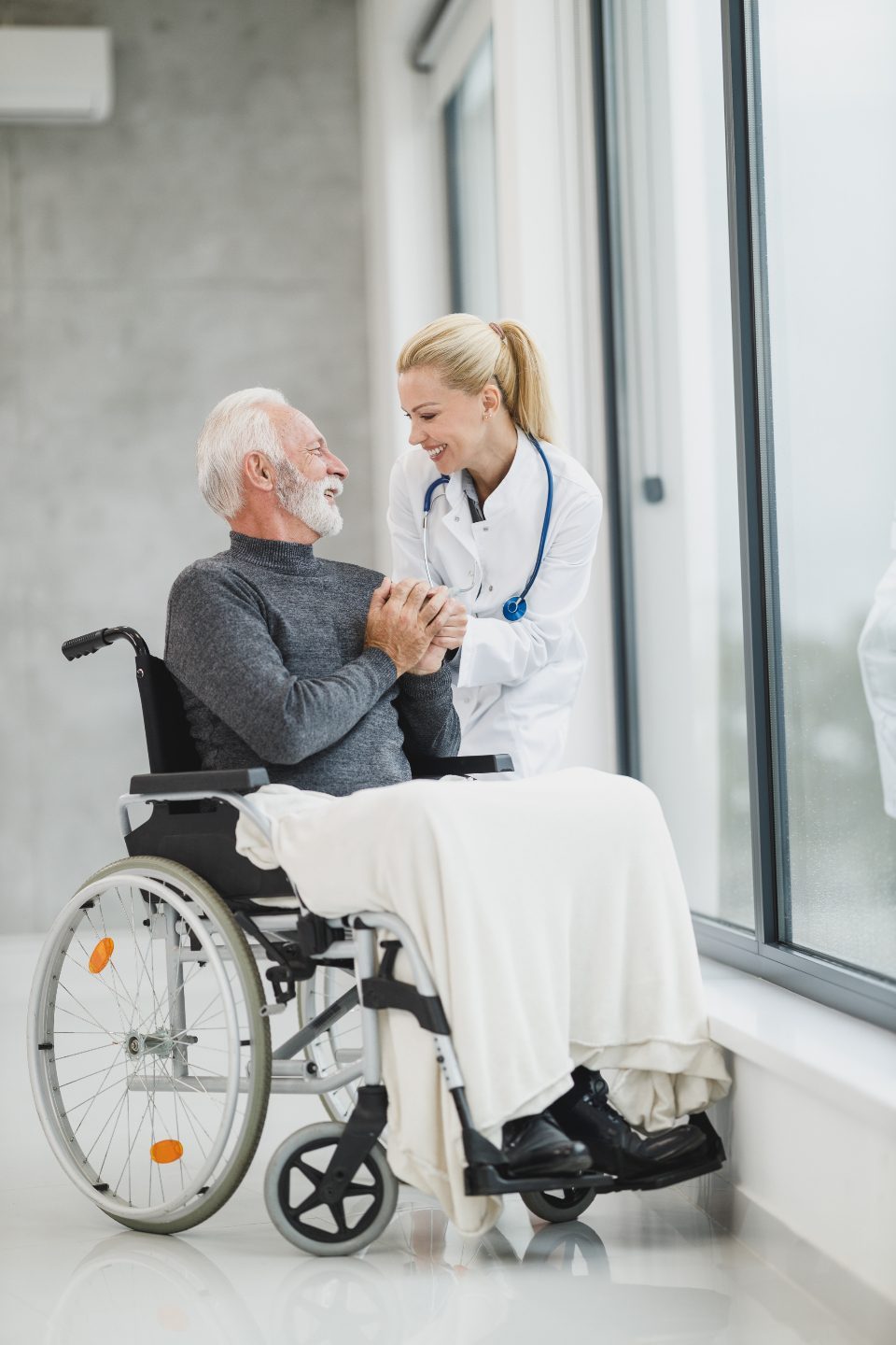 A clinician holding the hand of an older man in a wheelchair by a window